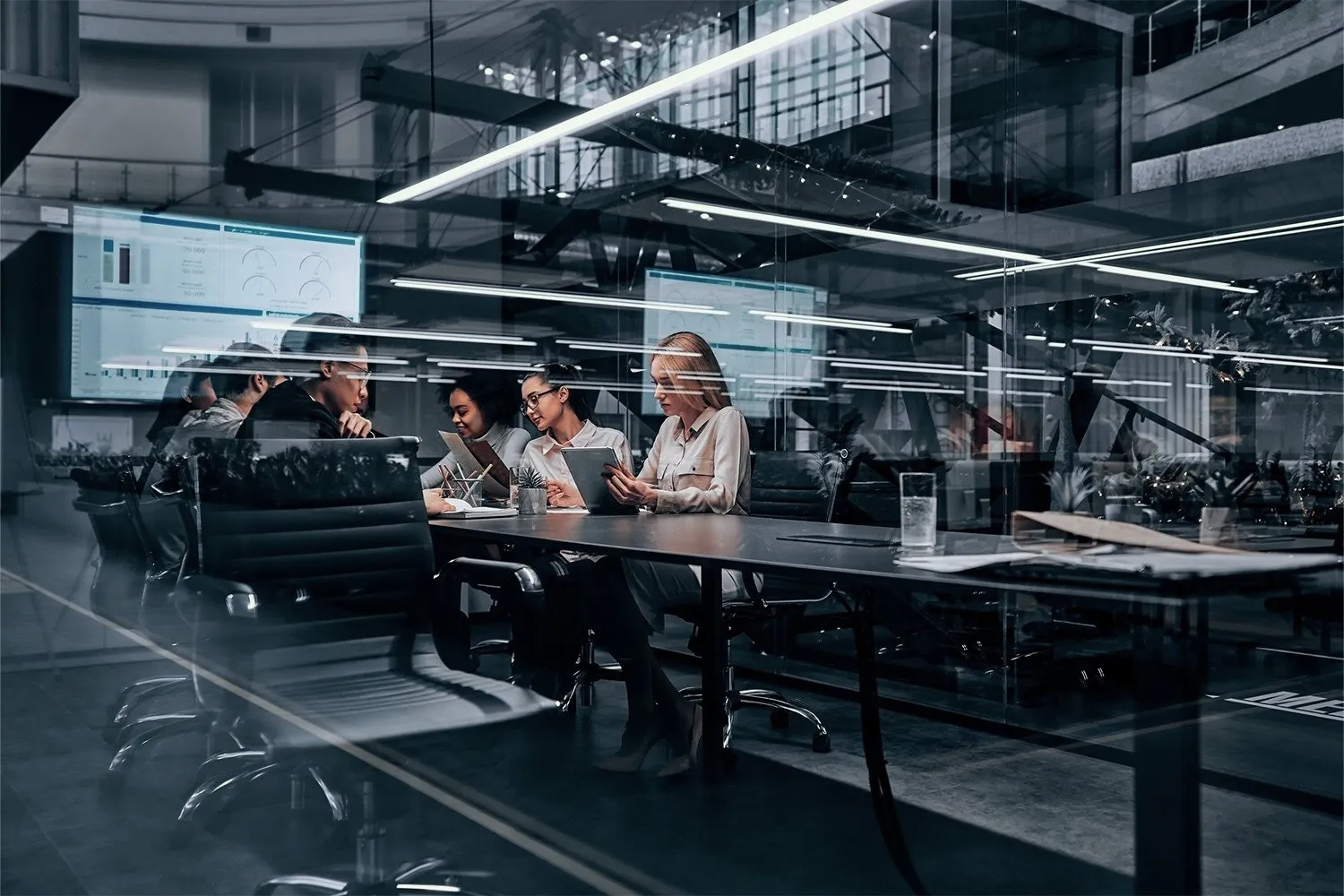People work at desks in a glass-walled operations room with screens along the wall.