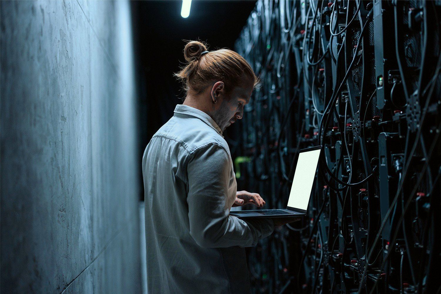 An engineer works on a laptop beside a row of dark server racks, cables running down the open cabinet.