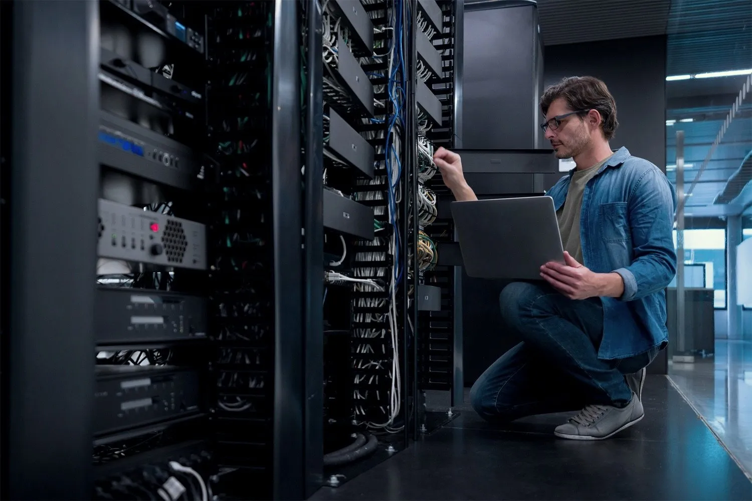 An engineer kneels at an open rack with a laptop on his knee, reaching in to a patch panel.