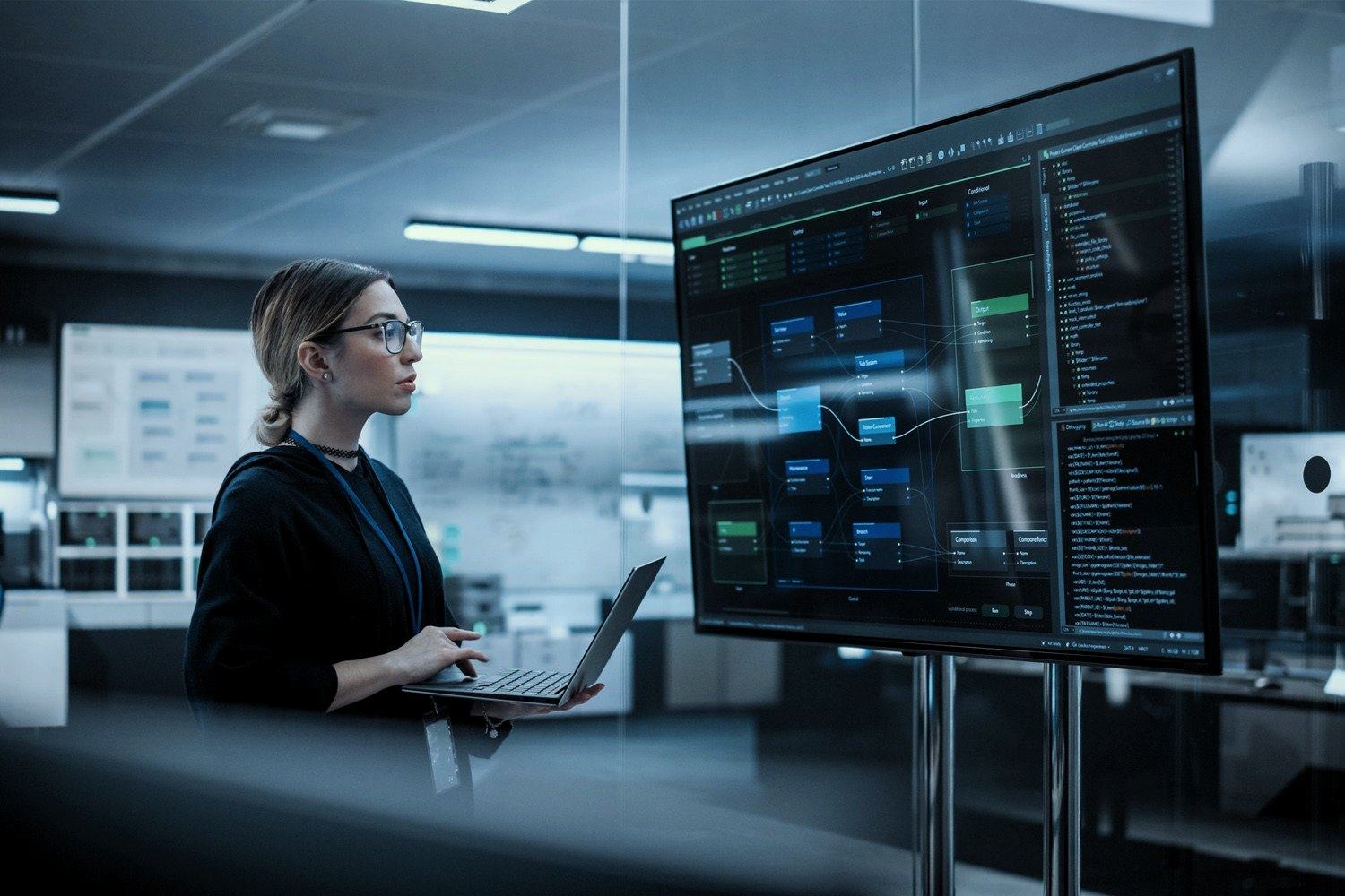 Stand-in photo: an engineer with a laptop studies a large monitoring screen in an operations room.