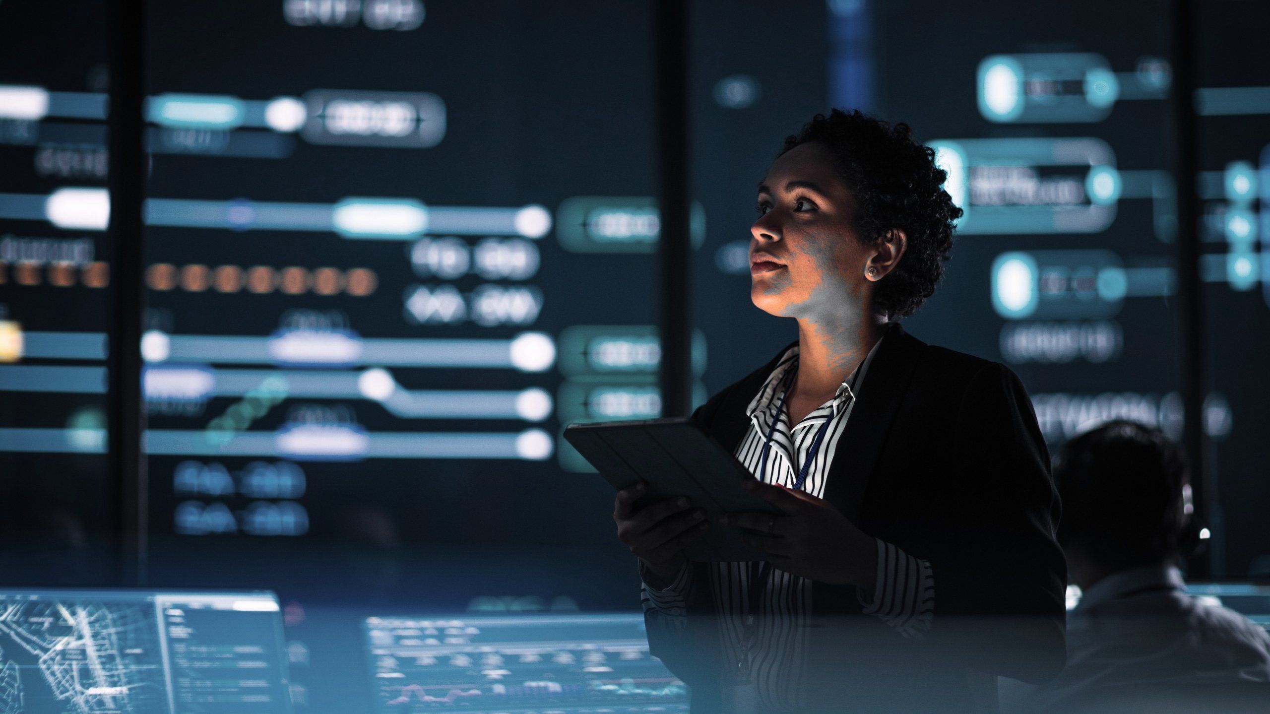 An engineer holding a tablet looks up at large monitoring screens in an operations centre.